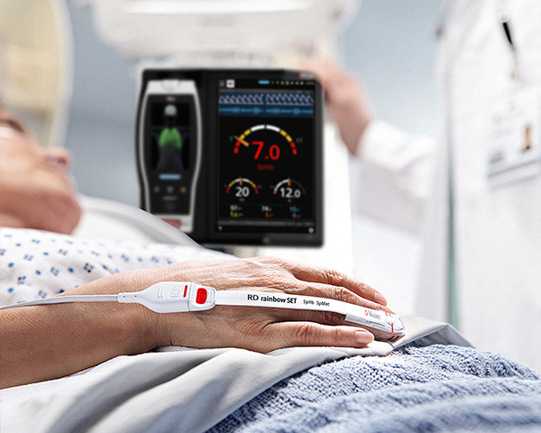 Patient in hospital bed monitored with Masimo sensor with root device and clinician in background checking on Root device.