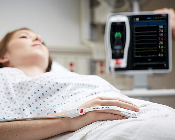 Patient sleeping in hospital bed wearing Masimo fingertip sensor with Root device in background.