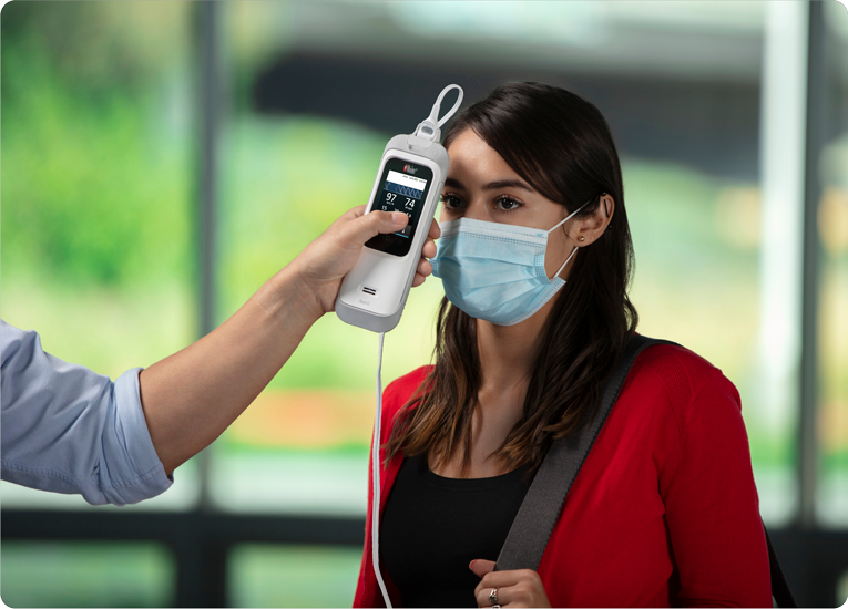 A healthcare provider checks a masked woman’s temperature using a Masimo Rad-G with Temperature device in an indoor setting.